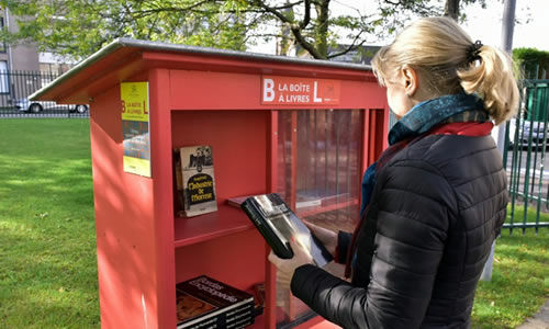 Une femme est vue de dos dans un parc avec un livre à la main devant une boite rouge contenant des livres