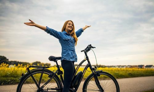 Une femme est sur un vélo noir devant un champs et elle lève les bras au ciel