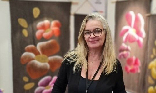 on voit une jeune femme blonde avec des lunettes : Virginie Soulier, fondatrice de Nigriv, avec des tableaux de fleurs en arrière plan. 