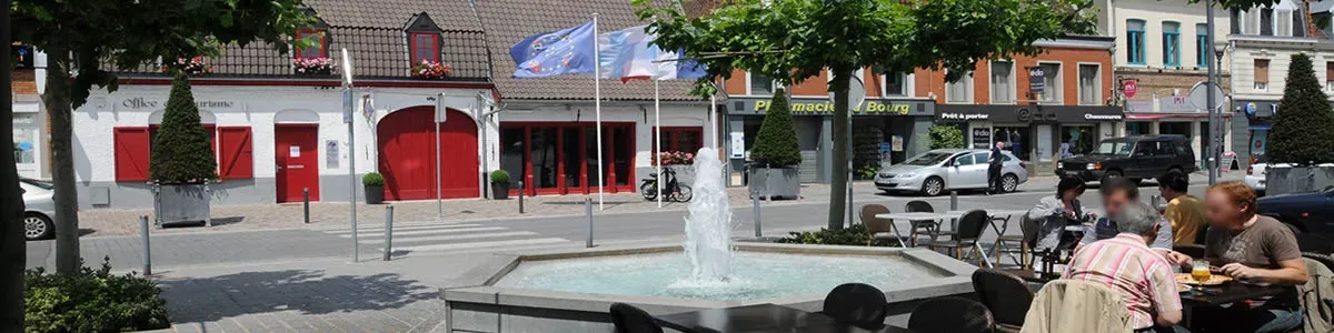 Une place avec une terrasse avec des personnes qui dejeunent devant une fontaine et la forge un bâtiment ancien aux volets et porte en bois rouges