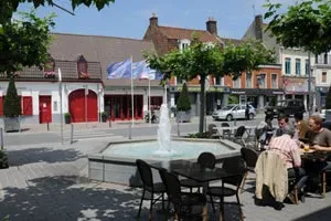 Une place une terrase avec des personnes qui dejeunent devant une fontaine et la forge un bâtiment ancien aux volets et porte en bois rouges