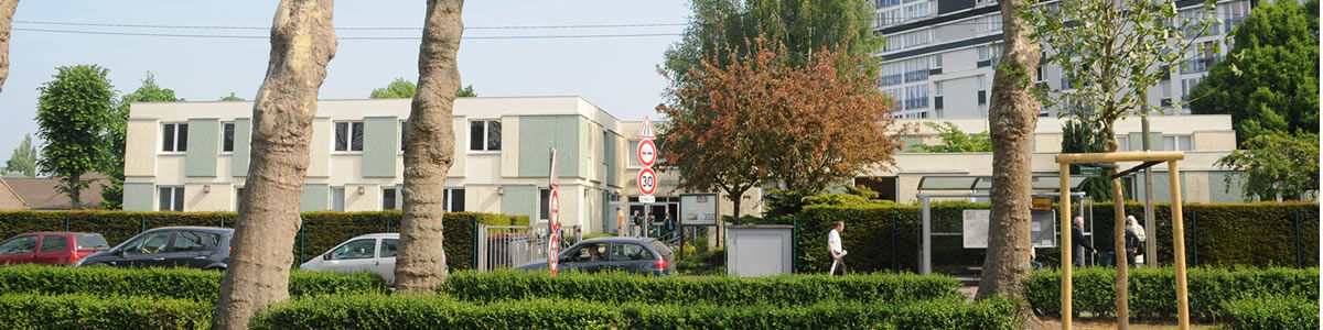 Le centre social un batimane bas blanc et vert avec premier plan une haie et des arbres, en fond à droite on aperçoit un bâtiment résidentiel de 9 étages